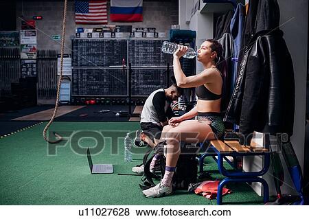 Stock Photo - Young woman training, sitting on bench drinking bottled water in gym. Fotosearch