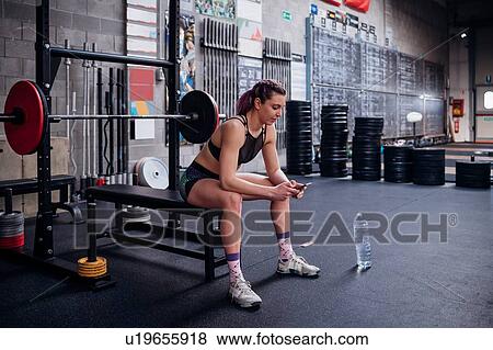Stock Photo - Young woman training, sitting on weights bench in gym looking at smartphone. Fotosearch