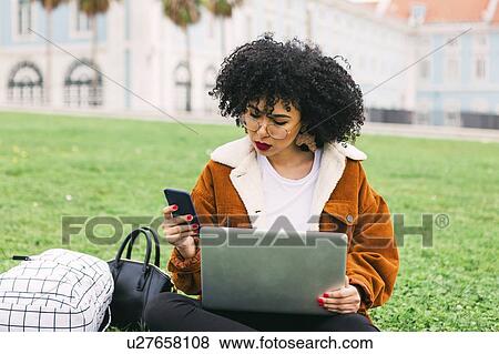 Stock Photo - Young woman using laptop and smart phone in park. Fotosearch