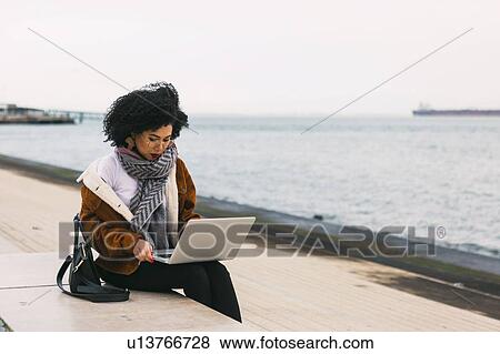 Stock Photo - Young woman using laptop on waterfront in Lisbon, Portugal. Fotosearch