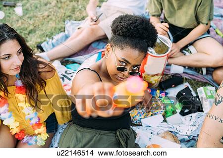 Picture - Young woman watching friend aiming water gun at picnic. Fotosearch