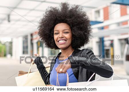 Young woman with afro hair at city train station holding up shopping bags, portrait View Large Photo Image Picture - Young woman with afro hair at city train station holding up shopping bags, portrait. Fotosearch