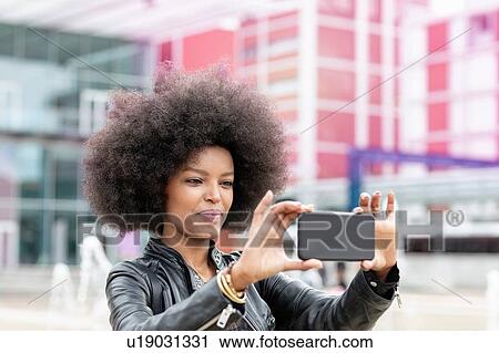 Stock Image - Young woman with afro hair at city train station, taking smartphone selfie. Fotosearch