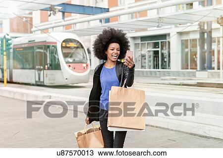 Stock Image - Young woman with afro hair at city train station carrying shopping bags, taking smartphone selfie. Fotosearch