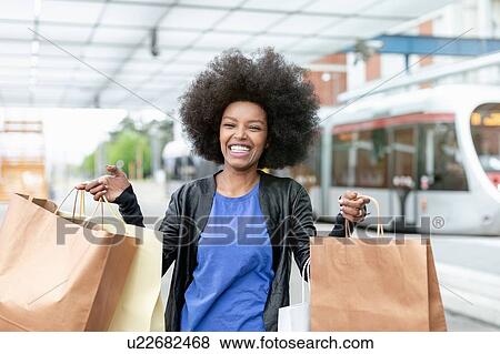 Stock Photo - Young woman with afro hair at city train station holding up shopping bags, portrait. Fotosearch