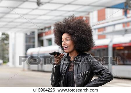 Stock Photograph - Young woman with afro hair at city train station, talking to smartphone. Fotosearch