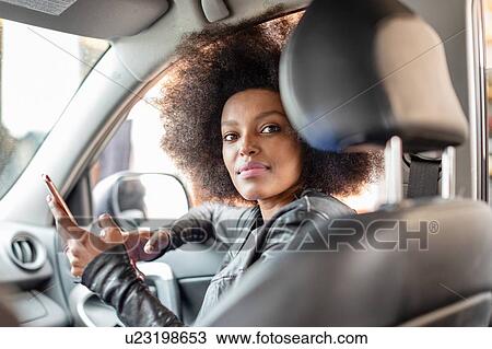 Stock Image - Young woman with afro hair in car passenger seat holding smartphone, portrait. Fotosearch