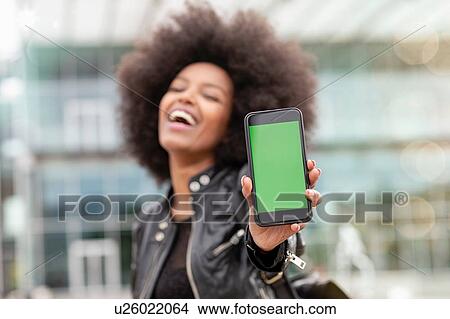 Young woman with afro hair in city, laughing and holding up smartphone View Large Photo Image Picture - Young woman with afro hair in city, laughing and holding up smartphone. Fotosearch