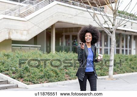 Stock Image - Young woman with afro hair in city, walking and talking to smartphone. Fotosearch