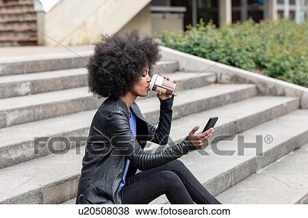 Stock Photo - Young woman with afro hair sitting on city stairway, drinking takeaway coffee and looking at smartphone. Fotosearch