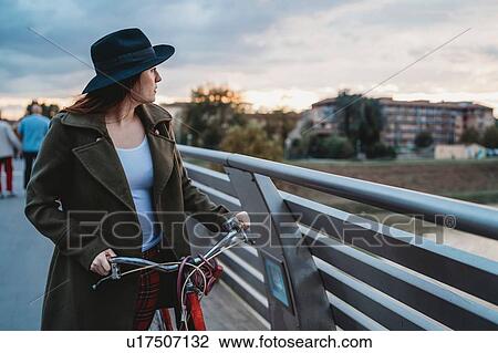 Stock Image - Young woman with bicycle looking out from footbridge at dusk, Florence, Tuscany, Italy. Fotosearch