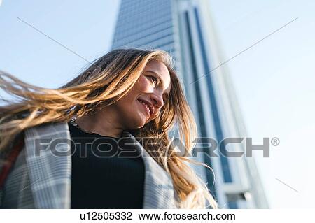Stock Image - Young woman with long blond hair in front of skyscraper, low angle view, Turin, Piemonte, Italy. Fotosearch