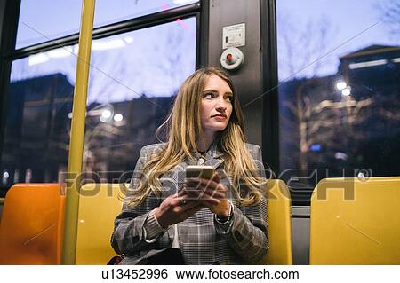 Stock Photograph - Young woman with long blond hair sitting in train carriage with smartphone at dusk. Fotosearch