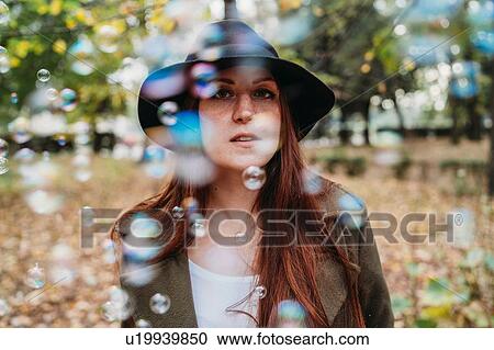 Stock Image - Young woman with long red hair amongst floating bubbles in autumn park, shallow focus portrait. Fotosearch