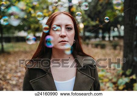 Stock Image - Young woman with long red hair amongst floating bubbles in autumn park, shallow focus portrait. Fotosearch