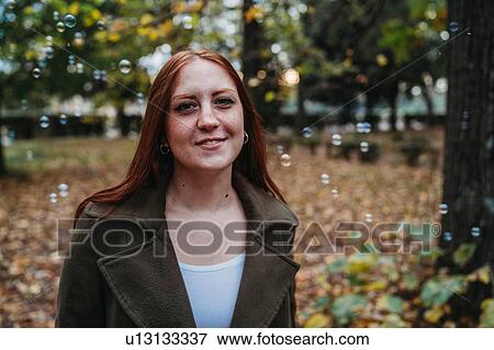 Stock Photo - Young woman with long red hair amongst floating bubbles in autumn park, head and shoulder portrait. Fotosearch