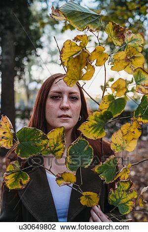 Stock Image - Young woman with long red hair behind twig of autumn leaves in park, portrait. Fotosearch