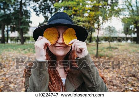 Stock Photo - Young woman with long red hair covering eyes with autumn leaves in park, portrait. Fotosearch