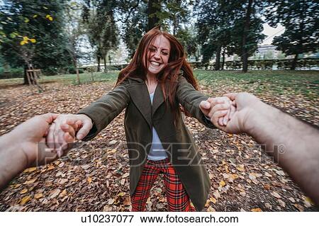 Stock Photo - Young woman with long red hair dancing while holding boyfriend's hands in autumn park, personal perspective. Fotosearch
