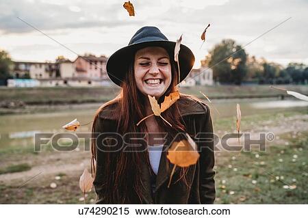 Stock Photography - Young woman with long red hair laughing amongst floating autumn leaves on riverside, Florence, Tuscany, Italy. Fotosearch