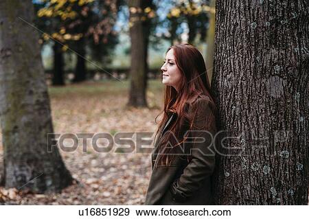 Young woman with long red hair leaning against tree trunk in autumn park View Large Photo Image Stock Photo - Young woman with long red hair leaning against tree trunk in autumn park. Fotosearch