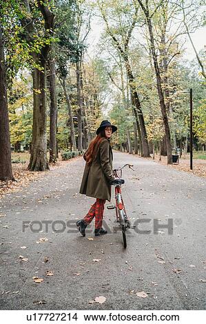 Picture - Young woman with long red hair looking over her shoulder while pushing bicycle in autumn park, full length, Florence, Tuscany, Italy. Fotosearch