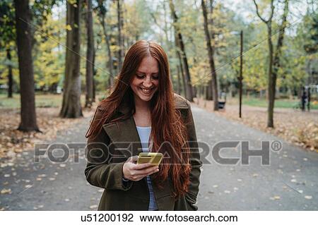 Stock Image - Young woman with long red hair looking at smartphone in tree lined autumn park, Florence, Tuscany, Italy. Fotosearch