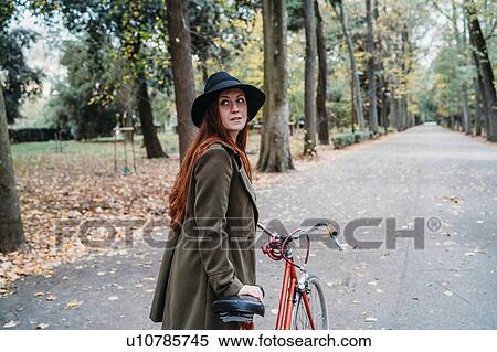 Stock Photography - Young woman with long red hair looking over her shoulder while pushing bicycle in autumn park, Florence, Tuscany, Italy. Fotosearch