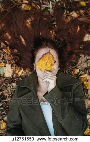 Stock Image - Young woman with long red hair lying amongst autumn leaves and covering face with autumn leaf, overhead portrait. Fotosearch