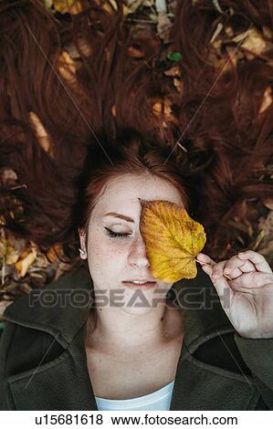 Stock Photo - Young woman with long red hair lying amongst autumn leaves and covering eye with autumn leaf, overhead portrait. Fotosearch