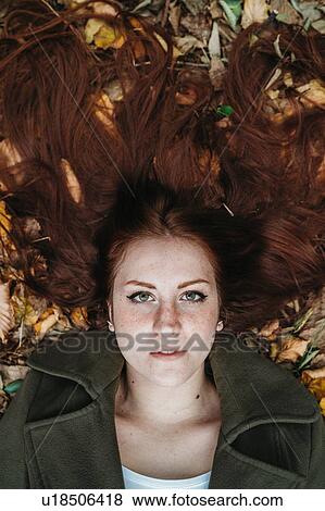 Stock Photo - Young woman with long red hair lying amongst autumn leaves, overhead portrait. Fotosearch