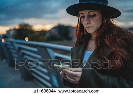 Picture - Young woman with long red hair on footbridge looking at smartphone at dusk, Florence, Tuscany, Italy. Fotosearch