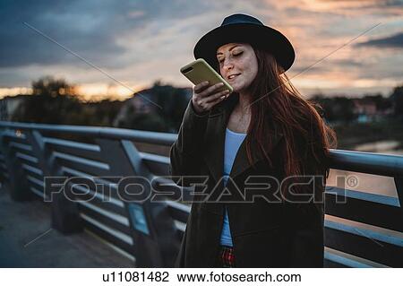 Stock Image - Young woman with long red hair on footbridge looking at smartphone at dusk, Florence, Tuscany, Italy. Fotosearch