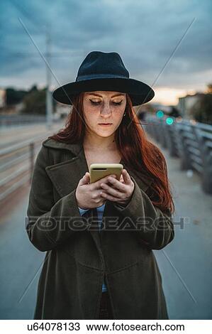 Stock Image - Young woman with long red hair on footbridge looking at smartphone at dusk. Fotosearch