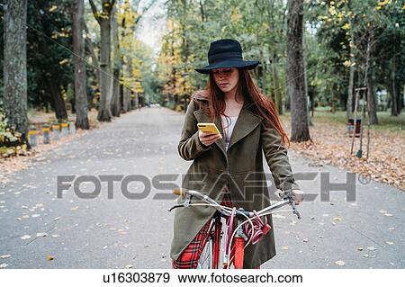 Stock Photo - Young woman with long red hair on bicycle looking at smartphone in autumn park, Florence, Tuscany, Italy. Fotosearch