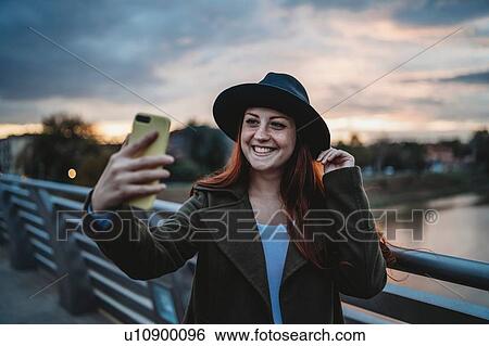 Stock Photograph - Young woman with long red hair on footbridge taking smartphone selfie at dusk, Florence, Tuscany, Italy. Fotosearch