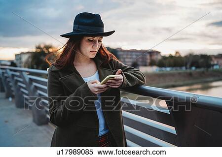 Stock Photography - Young woman with long red hair on footbridge looking at smartphone at dusk, Florence, Tuscany, Italy. Fotosearch