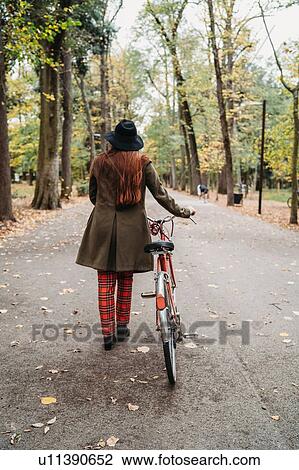 Stock Image - Young woman with long red hair pushing bicycle in autumn park, rear view full length, Florence, Tuscany, Italy. Fotosearch