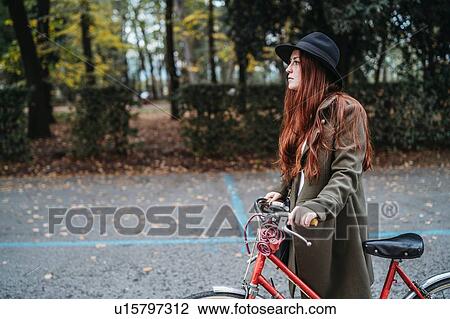 Stock Image - Young woman with long red hair pushing bicycle in autumn park. Fotosearch