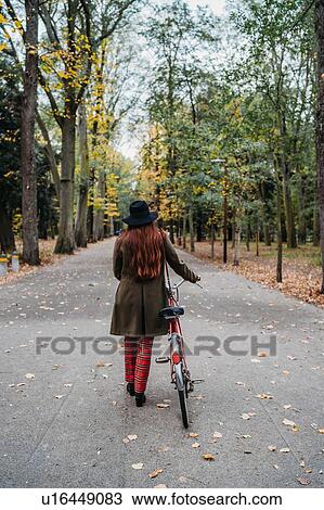 Stock Image - Young woman with long red hair pushing bicycle in tree lined autumn park, rear view, Florence, Tuscany, Italy. Fotosearch