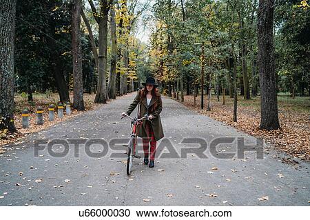 Stock Image - Young woman with long red hair pushing bicycle in tree lined autumn park, Florence, Tuscany, Italy. Fotosearch