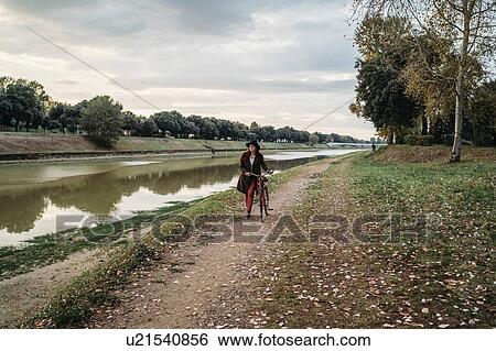 Stock Photograph - Young woman with long red hair pushing bicycle on riverside, full length, Florence, Tuscany, Italy. Fotosearch