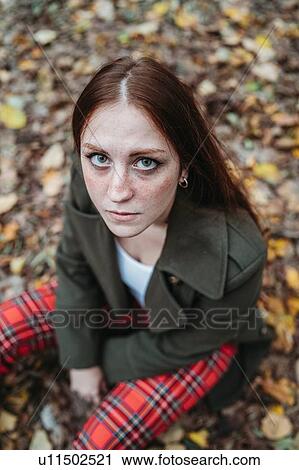 Stock Image - Young woman with long red hair sitting on autumn leaves in park, high angle portrait. Fotosearch