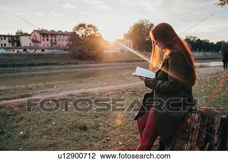 Stock Image - Young woman with long red hair sitting on tree stump reading book on riverside at sunset, Florence, Tuscany, Italy. Fotosearch