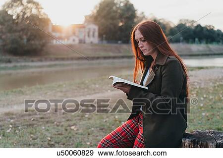 Stock Photo - Young woman with long red hair sitting on tree stump reading book on riverside at sunset, Florence, Tuscany, Italy. Fotosearch