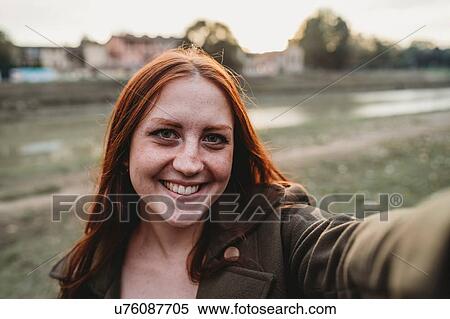 Stock Photography - Young woman with long red hair smiling for selfie on riverside, personal perspective, Florence, Tuscany, Italy. Fotosearch