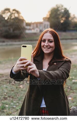 Stock Image - Young woman with long red hair taking smartphone selfie on riverside at dusk, Florence, Tuscany, Italy. Fotosearch