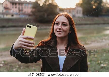 Stock Image - Young woman with long red hair taking smartphone selfie on riverside at dusk, Florence, Tuscany, Italy. Fotosearch