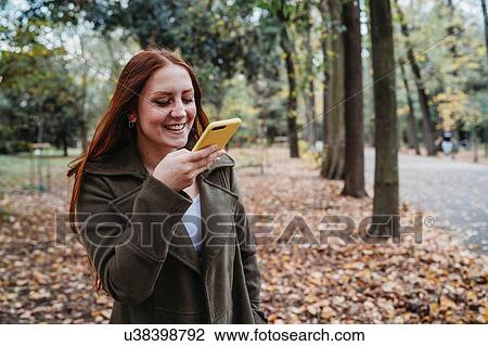 Stock Image - Young woman with long red hair taking smartphone selfie in autumn park. Fotosearch