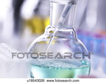 Stock Photograph - Chemistry research. Scientist taking a sample from a laboratory flask containing a chemical formula with a pipette.. Fotosearch